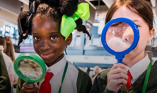 Three schoolchildren in uniform smiling and looking through colourful magnifying glasses indoors.