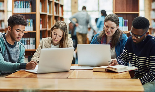 Four students working together at a library table with laptops, notebooks, and textbooks, surrounded by bookshelves.