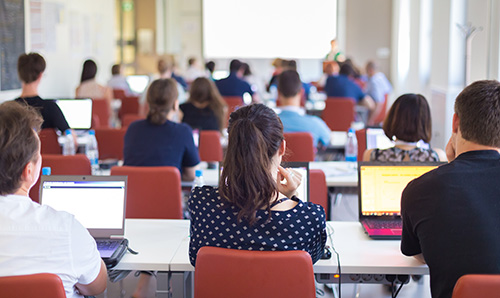 Students sitting in a classroom with laptops open, facing a projector screen during a lecture or training session.